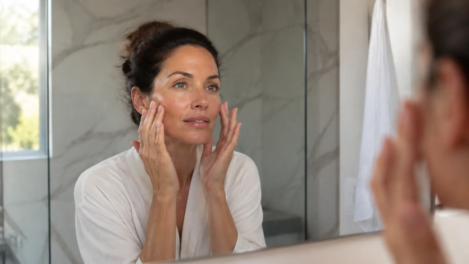 Woman applying serum at a bathroom mirror in the morning, skin looking healthy and comfortable