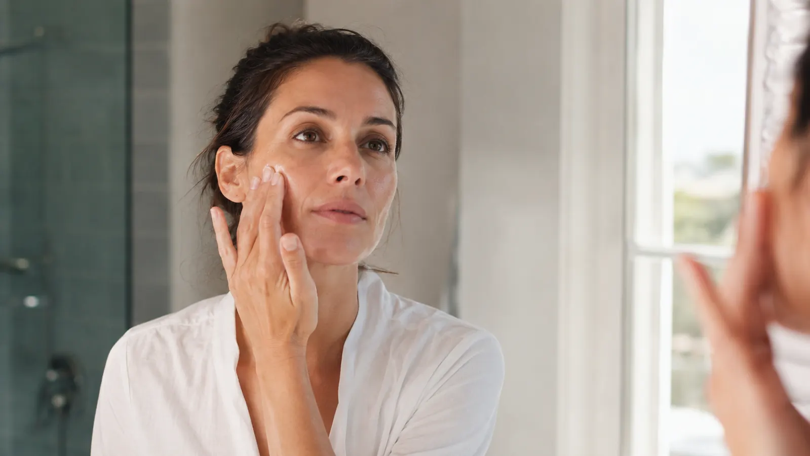 Woman applying serum to her cheek in a bright bathroom mirror, relaxed morning routine