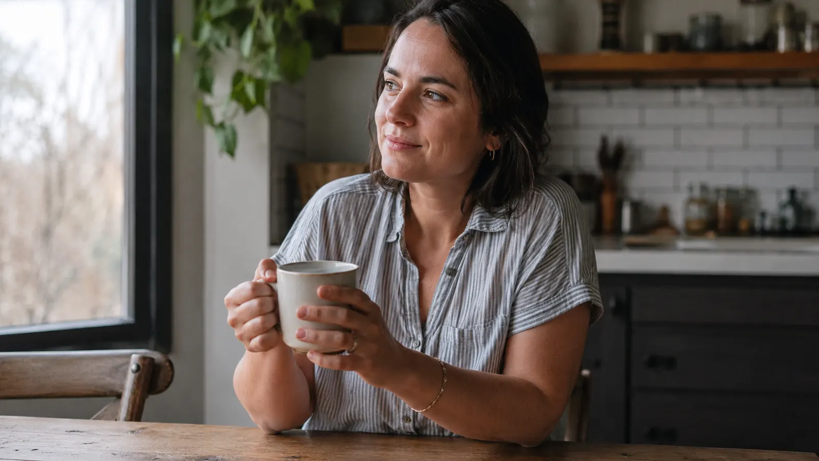 Woman in kitchen holding a coffee mug, looking relaxed and candid, morning light, no makeup