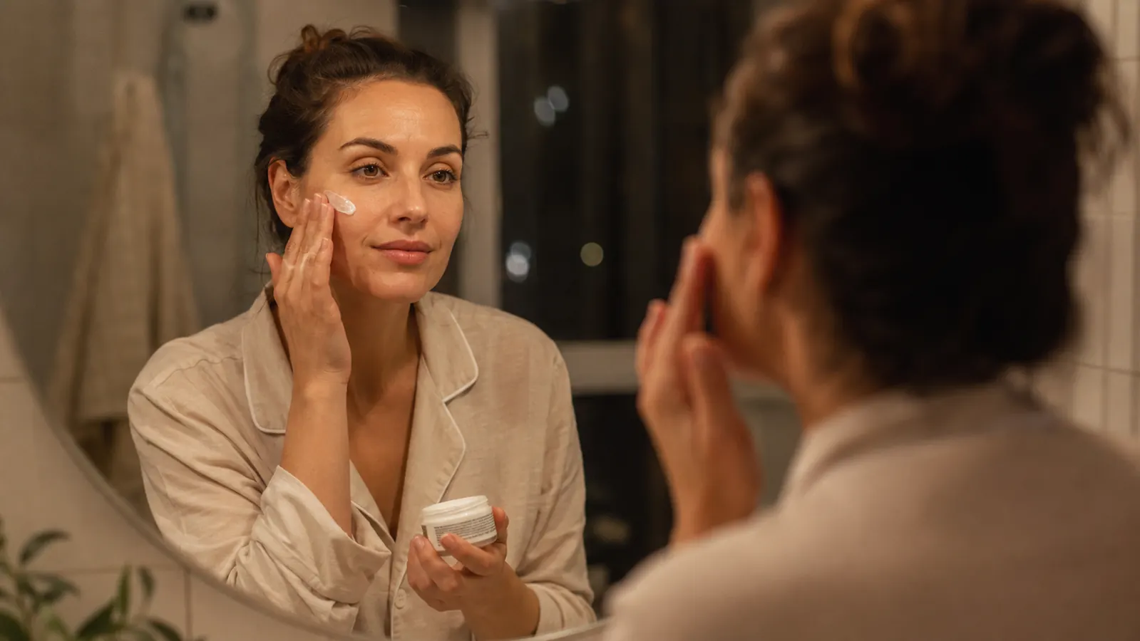 Woman with healthy, calm skin applying nighttime moisturizer in a bathroom mirror