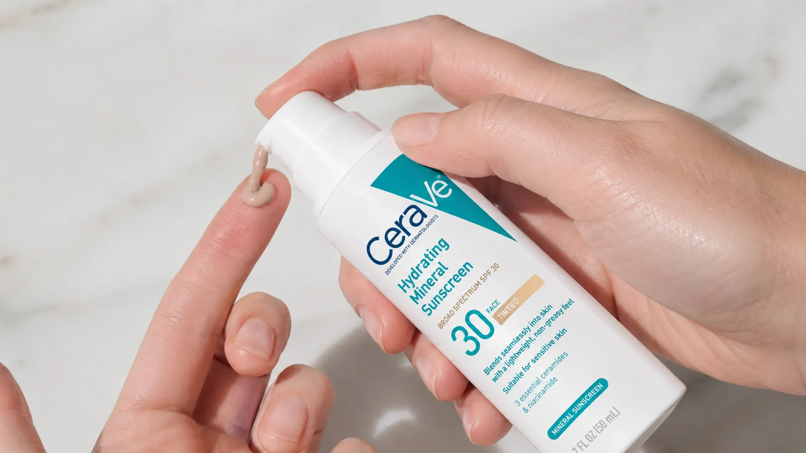 Hand holding the CeraVe Tinted Mineral Sunscreen tube, pump depressed, showing the beige-tinted product dispense