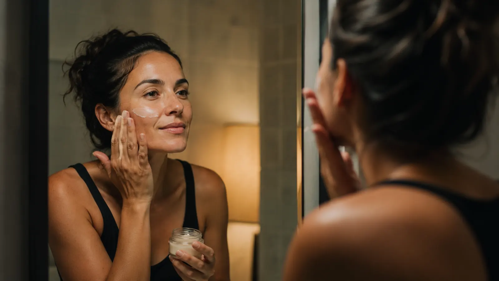 Woman patting moisturizer gently onto her cheeks at a bathroom mirror, calm expression, nighttime lighting