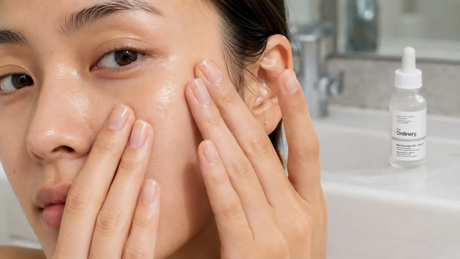 Hands pressing a few drops of clear serum into damp skin at a bathroom sink