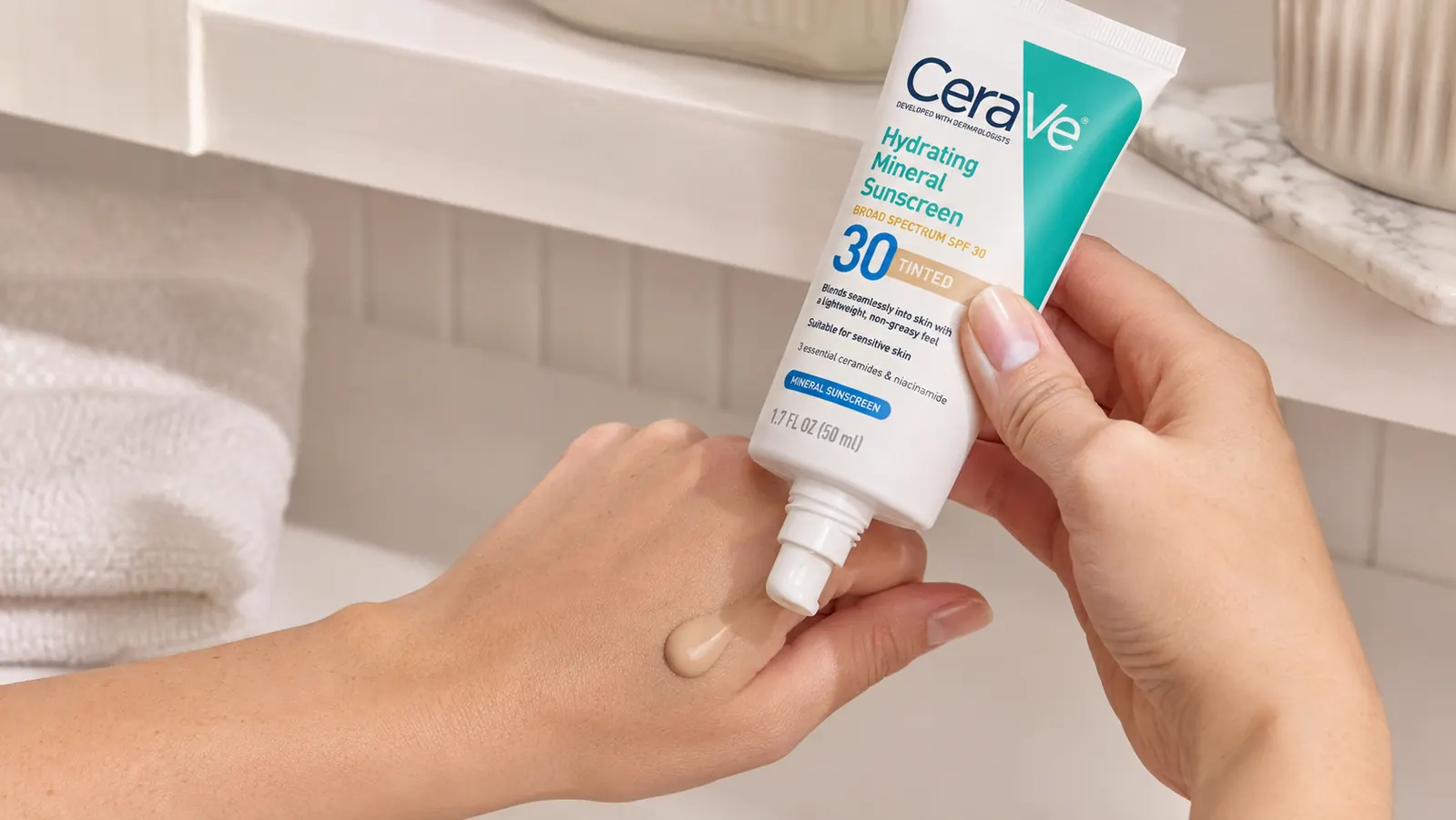 CeraVe Hydrating Mineral Sunscreen SPF 30 Tinted tube held in a palm next to moisturizer on a bathroom shelf