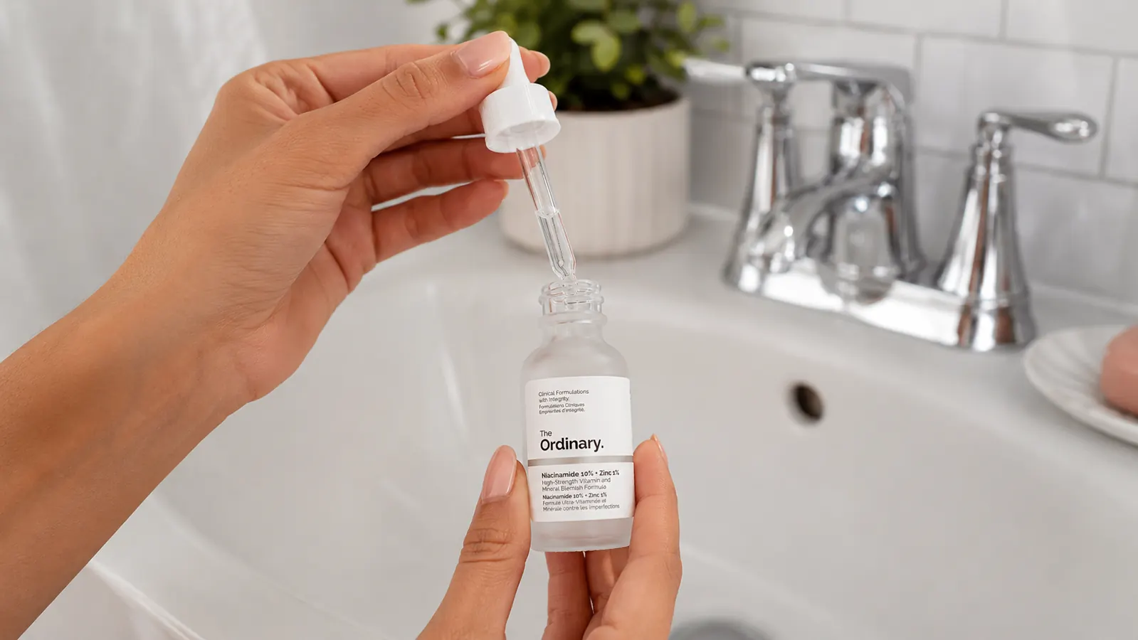 Hand holding The Ordinary Niacinamide serum bottle over a bathroom sink, cap removed, ready to dispense