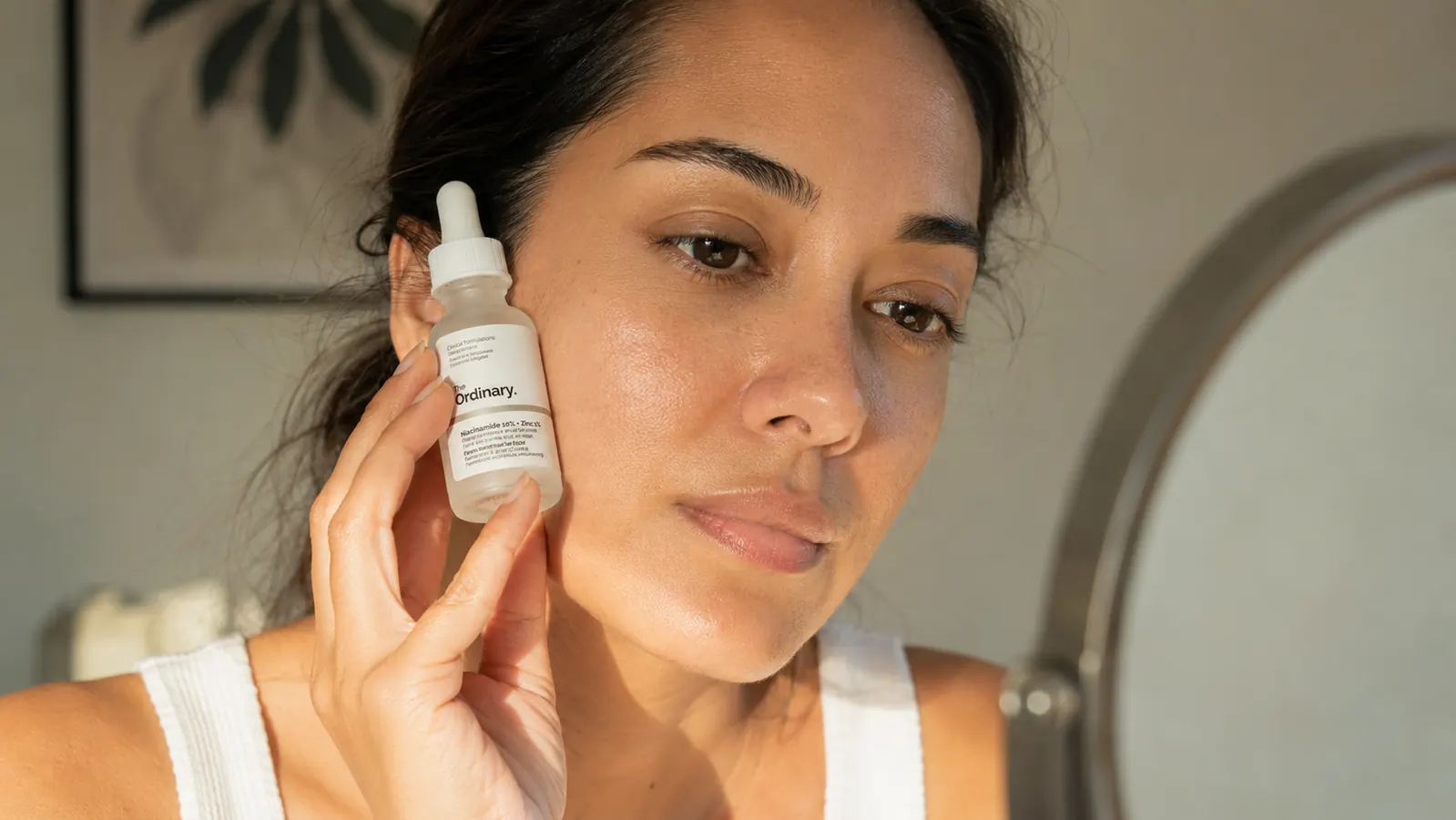 A close-up of a woman pressing a serum dropper gently to her cheek while looking in a bathroom mirror