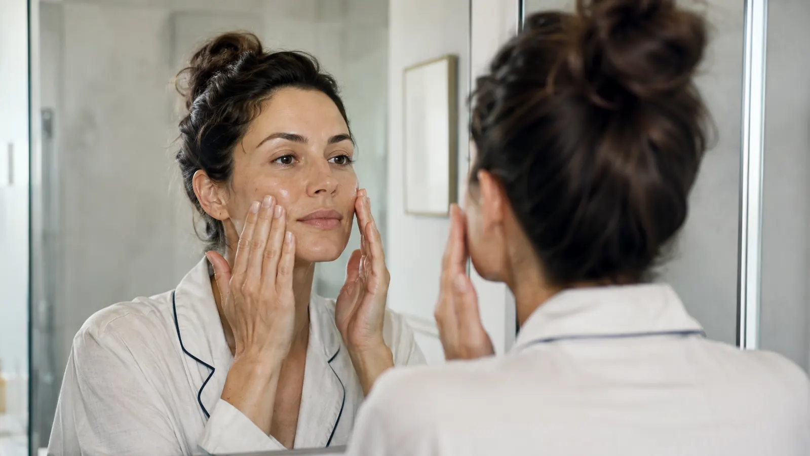 Woman patting serum into her cheek in front of a bathroom mirror, skin visibly calm