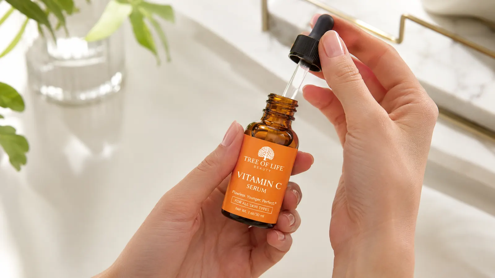 Hand holding a small amber serum bottle over a white bathroom countertop with a few skincare products nearby