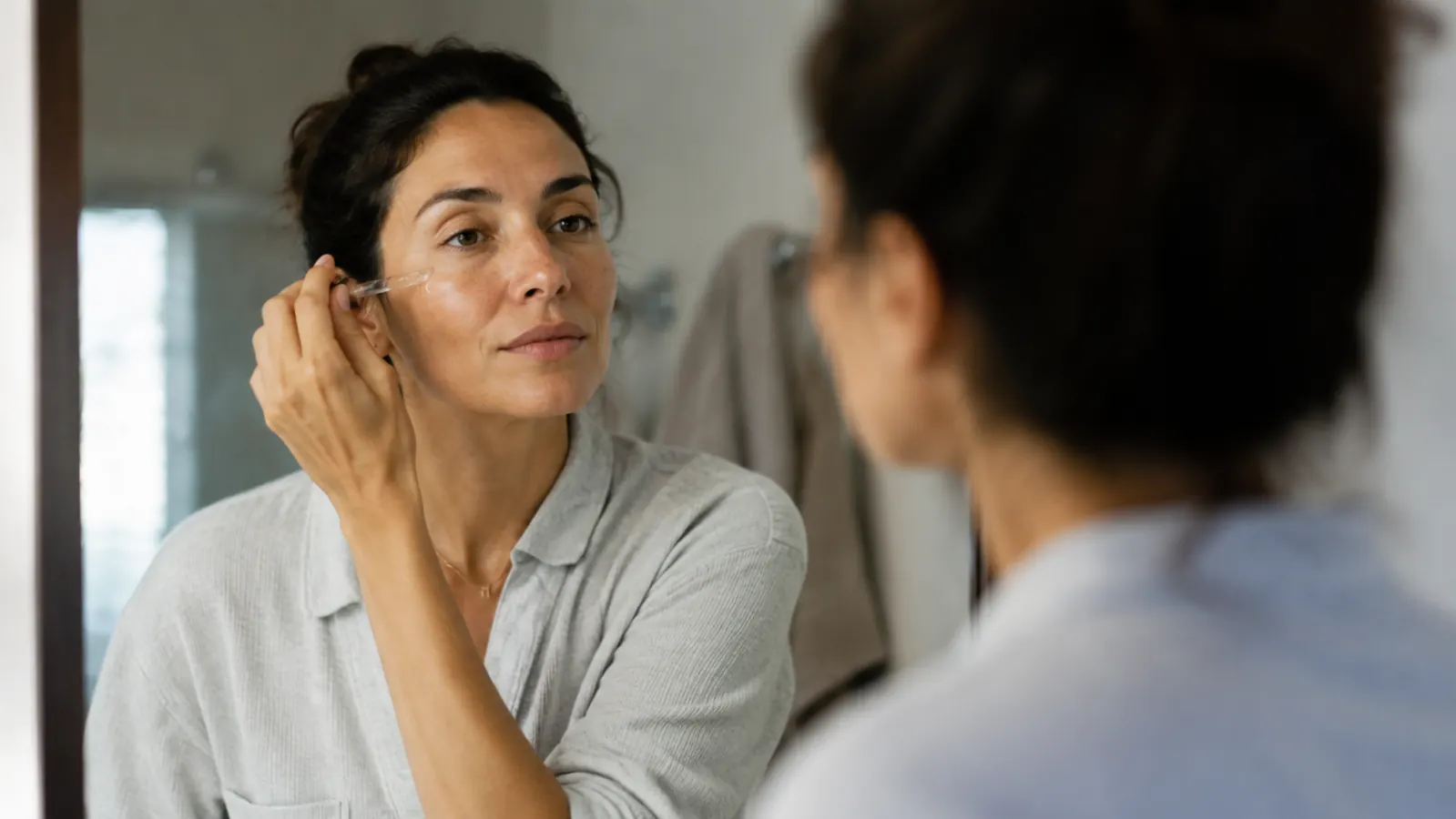 Woman with clear even skin applying a few drops of serum in front of a bathroom mirror in natural light